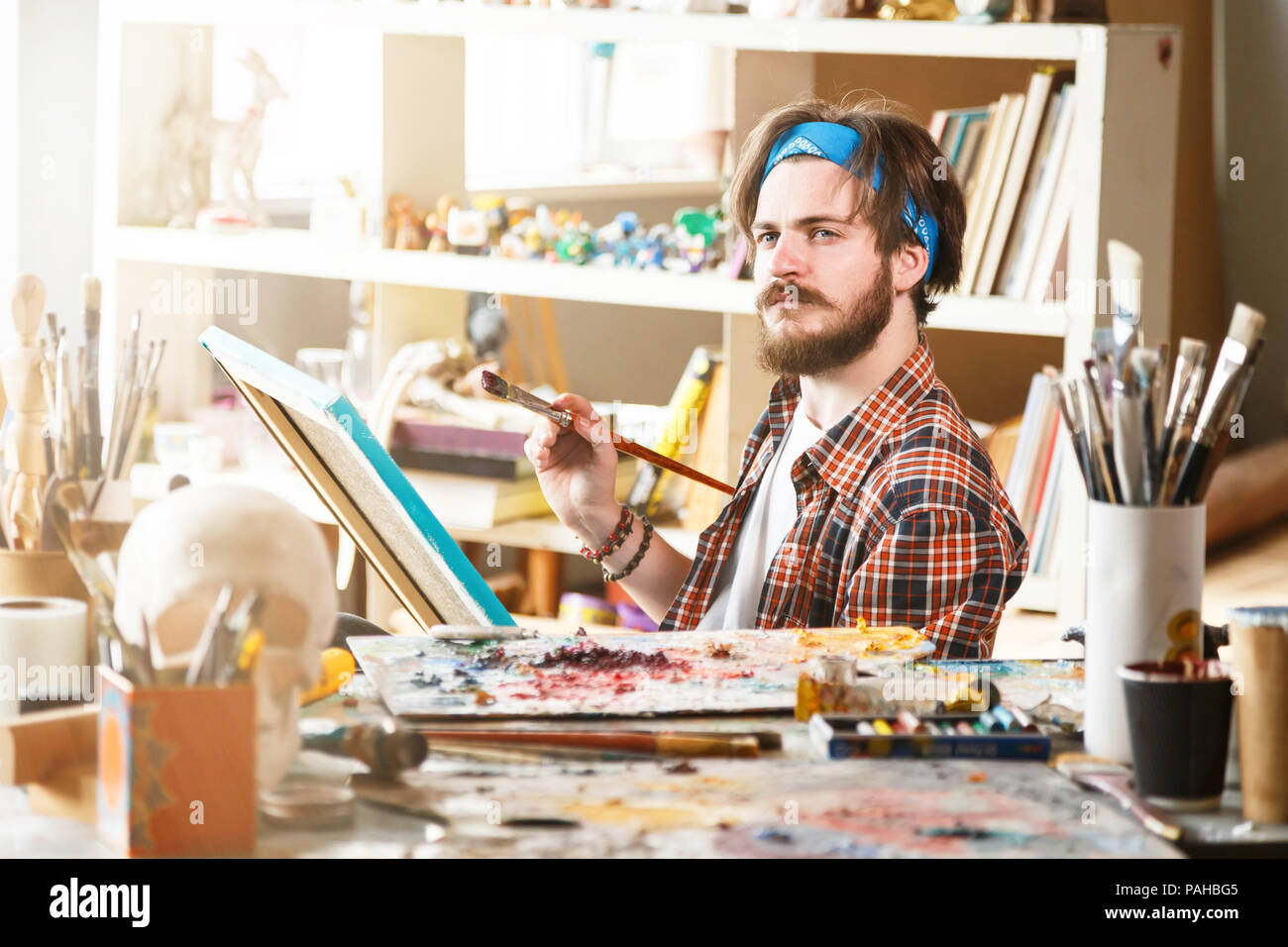 Young dark-haired caucasian male artist in blue bandana sitting and holding brush in his sunny studio Stock Photo