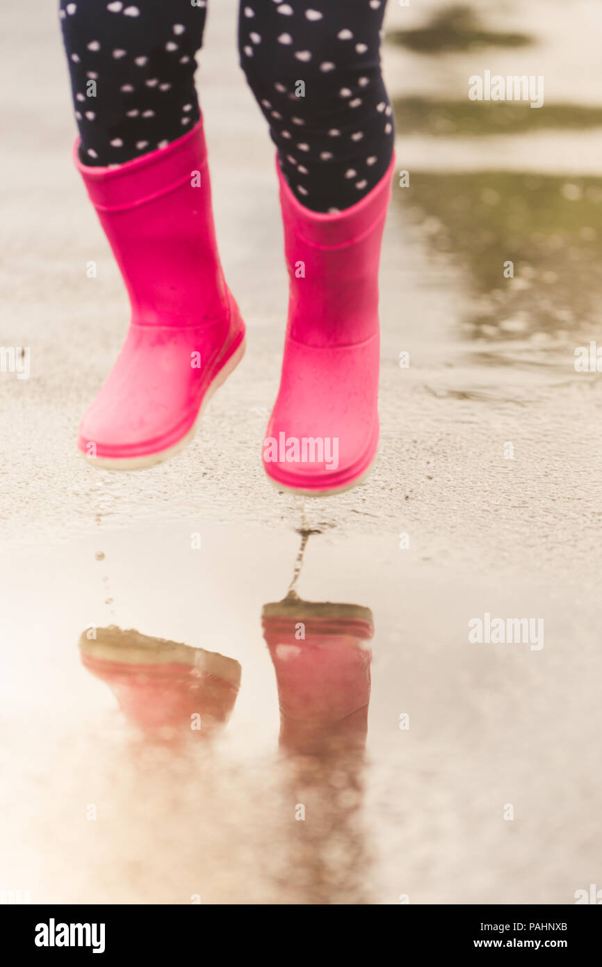 Feet of child in pink rubber boots jumping and splashing over puddle after rain. Stock Photo