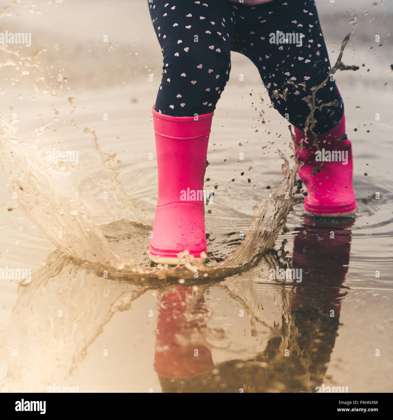 Feet of child in pink rubber boots jumping and splashing over puddle after rain. Stock Photo