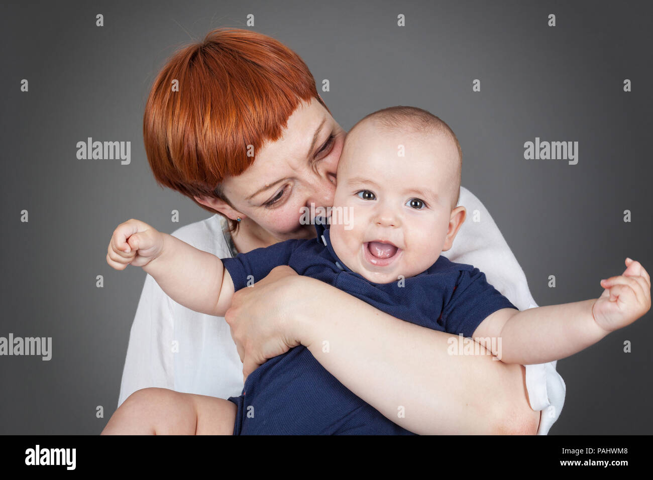Happy baby-Close up of Mother holding and kissing a joyfull baby boy who is laughing with both arms stretched out, isolated on grey with vignette, hor Stock Photo