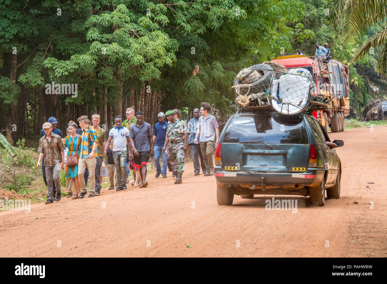 Group of students and tour guides walk together on dirt road as a car  packed with various household items drives past, Republic of Guinea Stock Photo