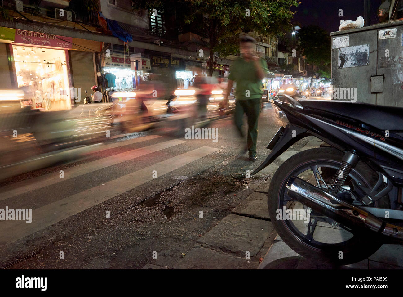 A motion blurred man walks next to busy traffic at night at the streets of Hanoi's Old Quarter, Vietnam, framed by a parked motorbike in the foregroun Stock Photo