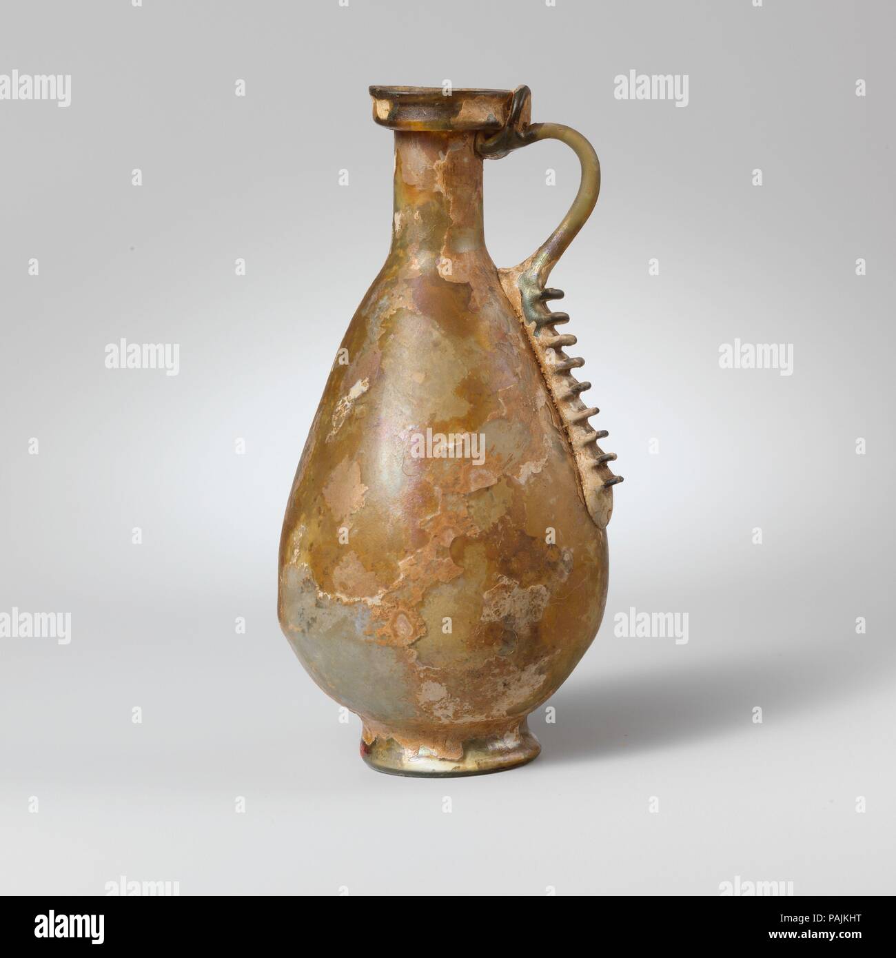 Glass jug. Culture: Roman. Dimensions: H.: 6 in. (15.2 cm). Date: mid-1st century A.D..  Colorless with pale blue green tinge; handle in same color.  Rim folded out, down, up, and out, forming a downward flange and stepped profile; short cylindrical neck with faint horizontal tooling marks around base; piriform body, with slight horizontal bulge at point of greatest diameter; low integral foot ring, concave bottom; rod handle attached to top of body with long tail extending down side and pinched to form nine semicircular projections, drawn up, out, and round in a curve, and trailed on to top o Stock Photo