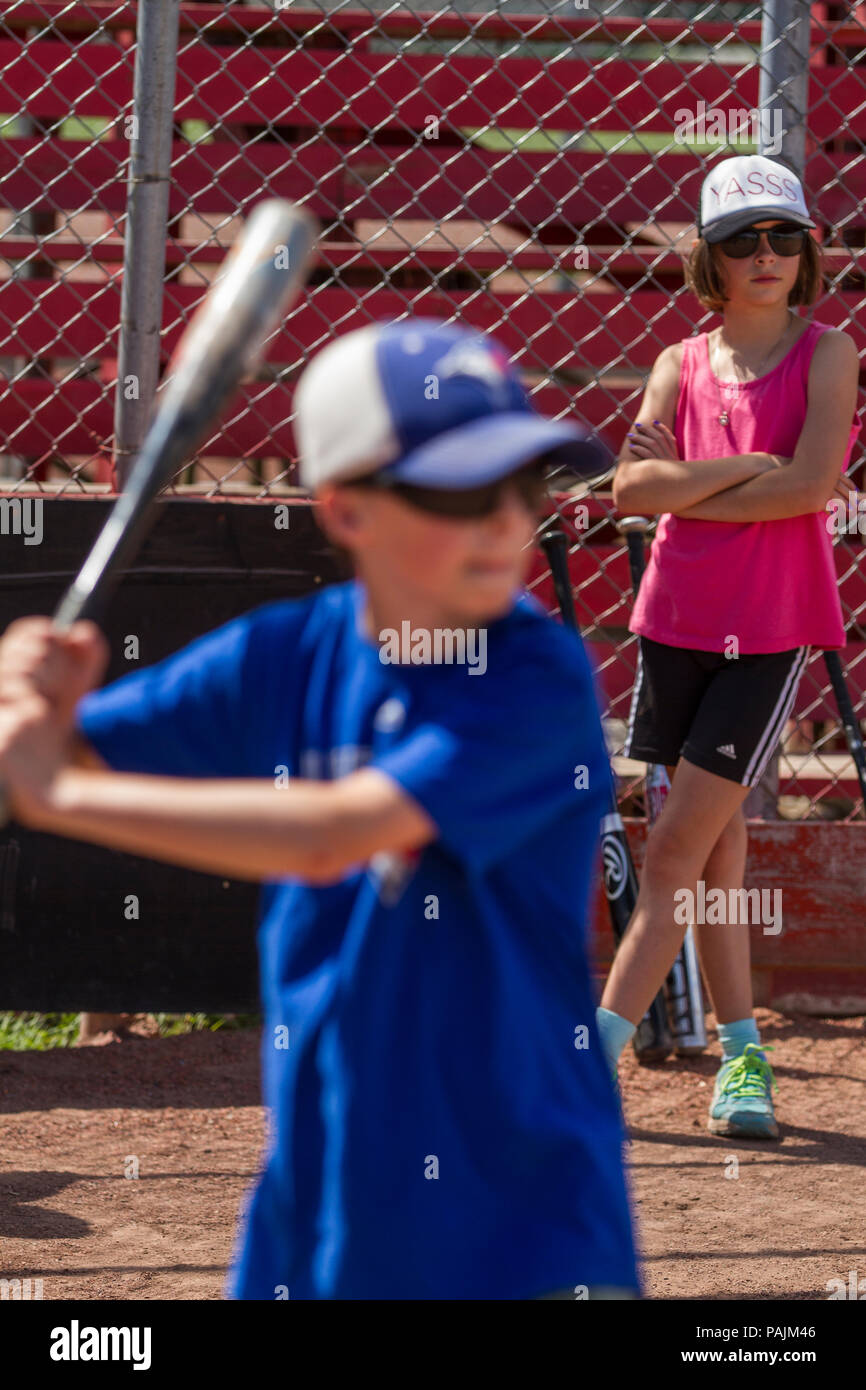 Red tank topped girl, in sunglasses, shorts and ball hat watching brother bat. Fore ground out of focus.  Model Relese $104 Girl #105 boy Stock Photo