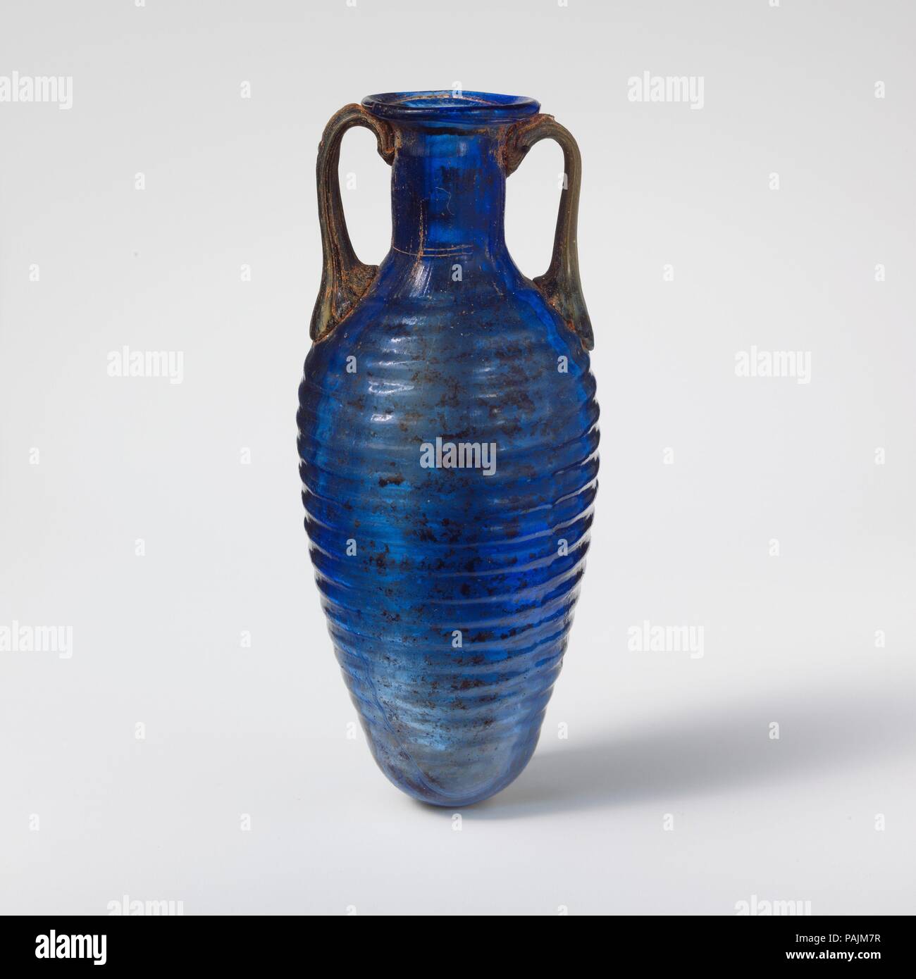 Glass amphoriskos with horizontal ribs. Culture: Roman. Dimensions: H. 4 5/16 in. (11 cm); diameter 1 13/16 in. (4.6 cm). Date: 2nd half of 1st century A.D..  Translucent cobalt blue, with handles in a semi-opaque mixture of blue, white, and yellowish brown.  Rim folded out, over, and in; flaring, uneven mouth; cylindrical neck with irregular horizontal ridges and indents around top; ovoid body, tapering to plain, rounded bottom; two rod handles attached to upper body in pads, drawn up and in, then pressed onto top of neck and underside of rim. One continuous mold seam around body, extending t Stock Photo