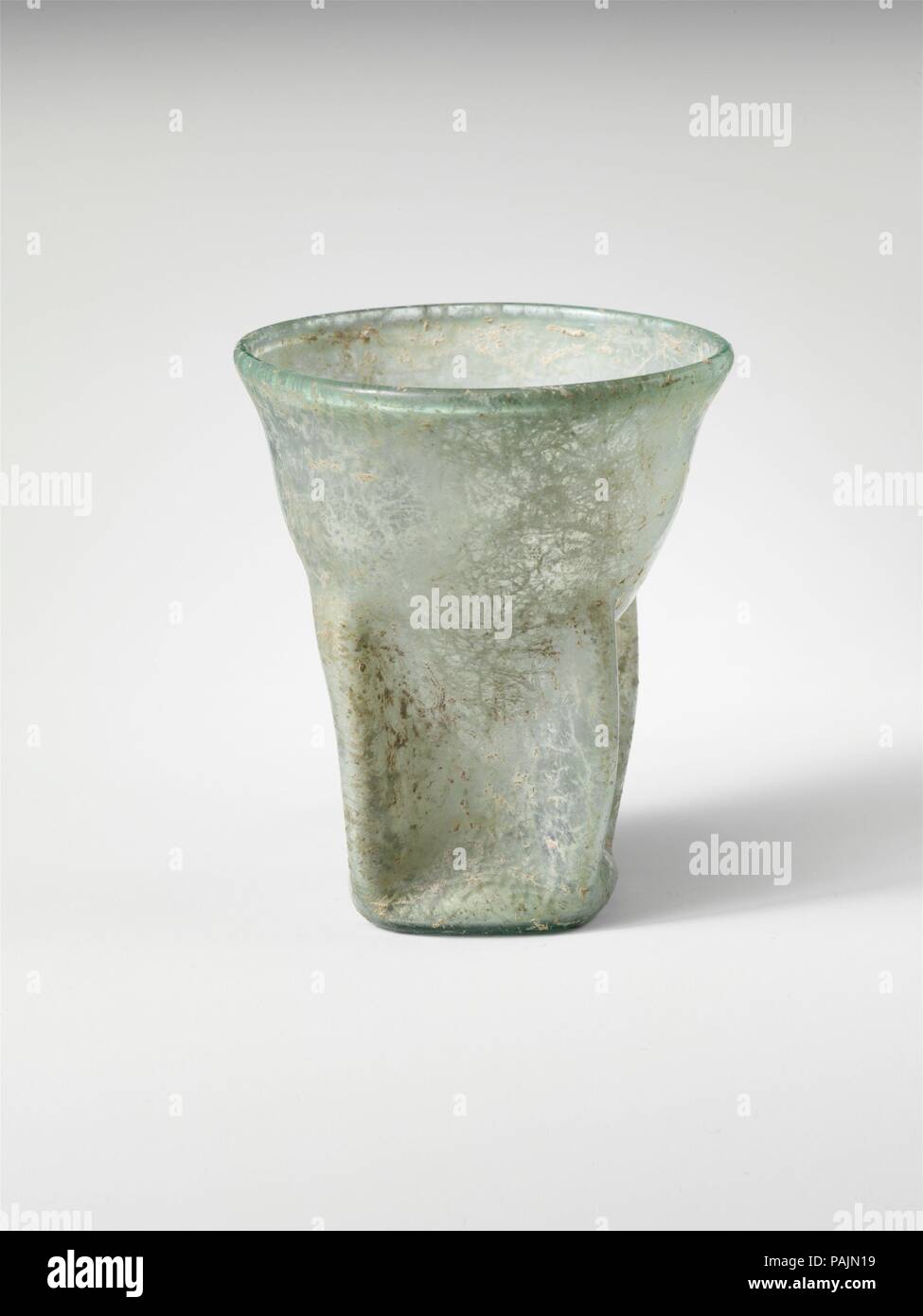 Glass beaker. Culture: Roman. Dimensions: H.: 3 1/2 x 3 in. (8.9 x 7.6 cm). Date: 3rd-4th century A.D..  Translucent blue green.  Thickened rim, with slightly inverted, rounded lip; side tapering downwards on upper body, and then shaped into a rough square with hollow projecting corners; roughly square, concave bottom.  On lower body, four deep indents, forming a star-like shape to interior.  Intact; few bubbles; soil encrustation and root marks covering most of surfaces. Museum: Metropolitan Museum of Art, New York, USA. Stock Photo
