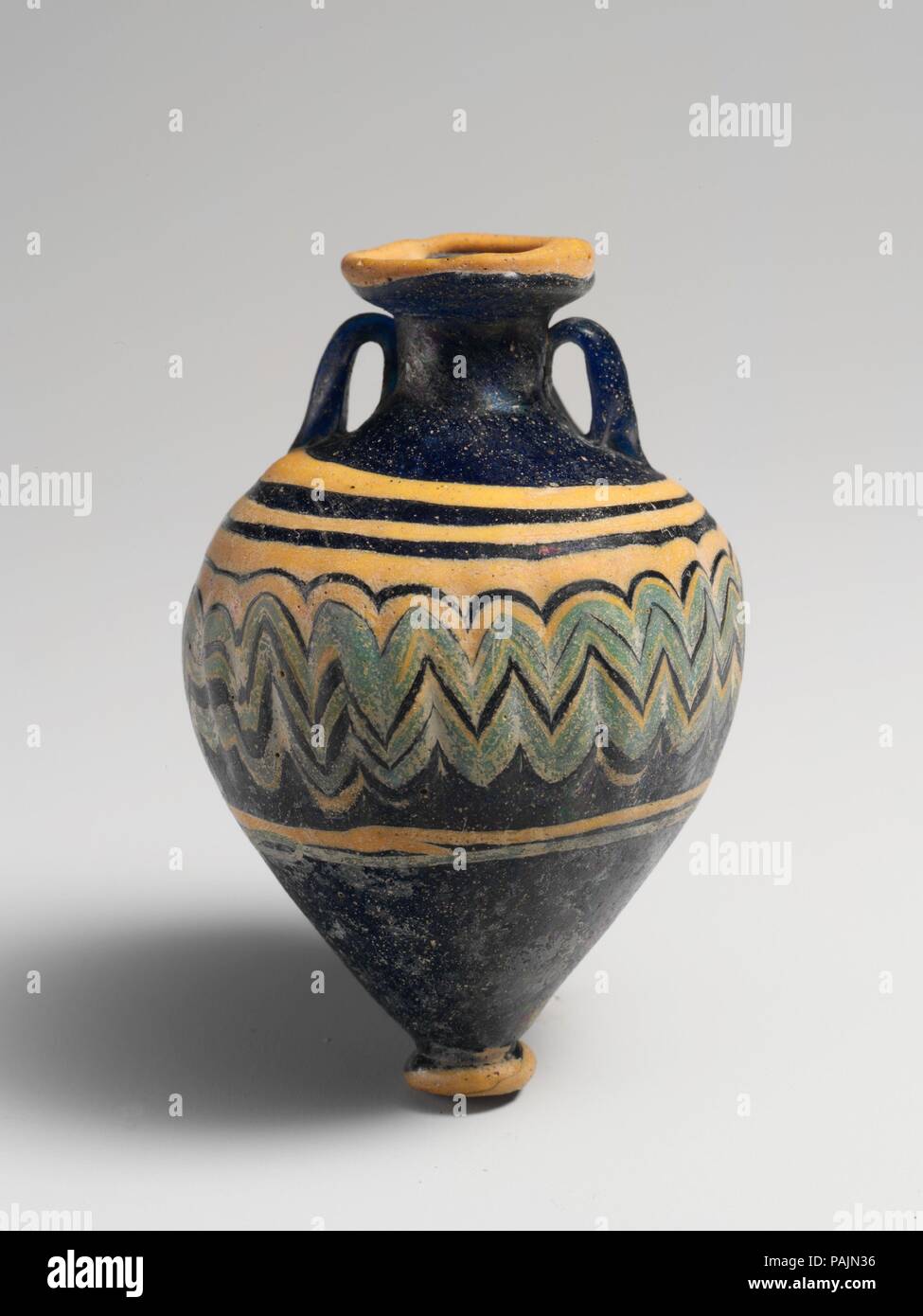 Glass amphoriskos (perfume bottle). Culture: Greek, Eastern Mediterranean. Dimensions: H.: 3 in. (7.5 cm). Date: late 6th-5th century B.C..  Translucent cobalt blue, with handles in same color; trails in opaque yellow and opaque turquoise blue.  Deeply inward-sloping rim-disk; cylindrical neck; broad sloping shoulder; top-shaped body; circular base-knob with flat bottom; two vertical strap handles applied to shoulder, drawn up and in, and pressed onto neck.  A thick yellow trail attached at edge of rim-disk; another thick yellow trail applied on shoulder, wound in a spiral, then tooled into an Stock Photo