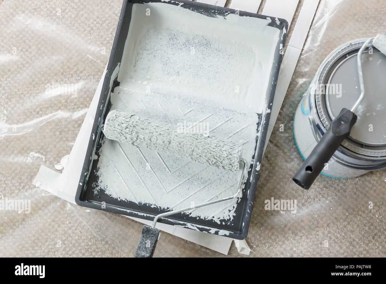 Painting and decorating with rollers and tray filled with light grey paint on a plastic dust sheet with cans in the background Stock Photo