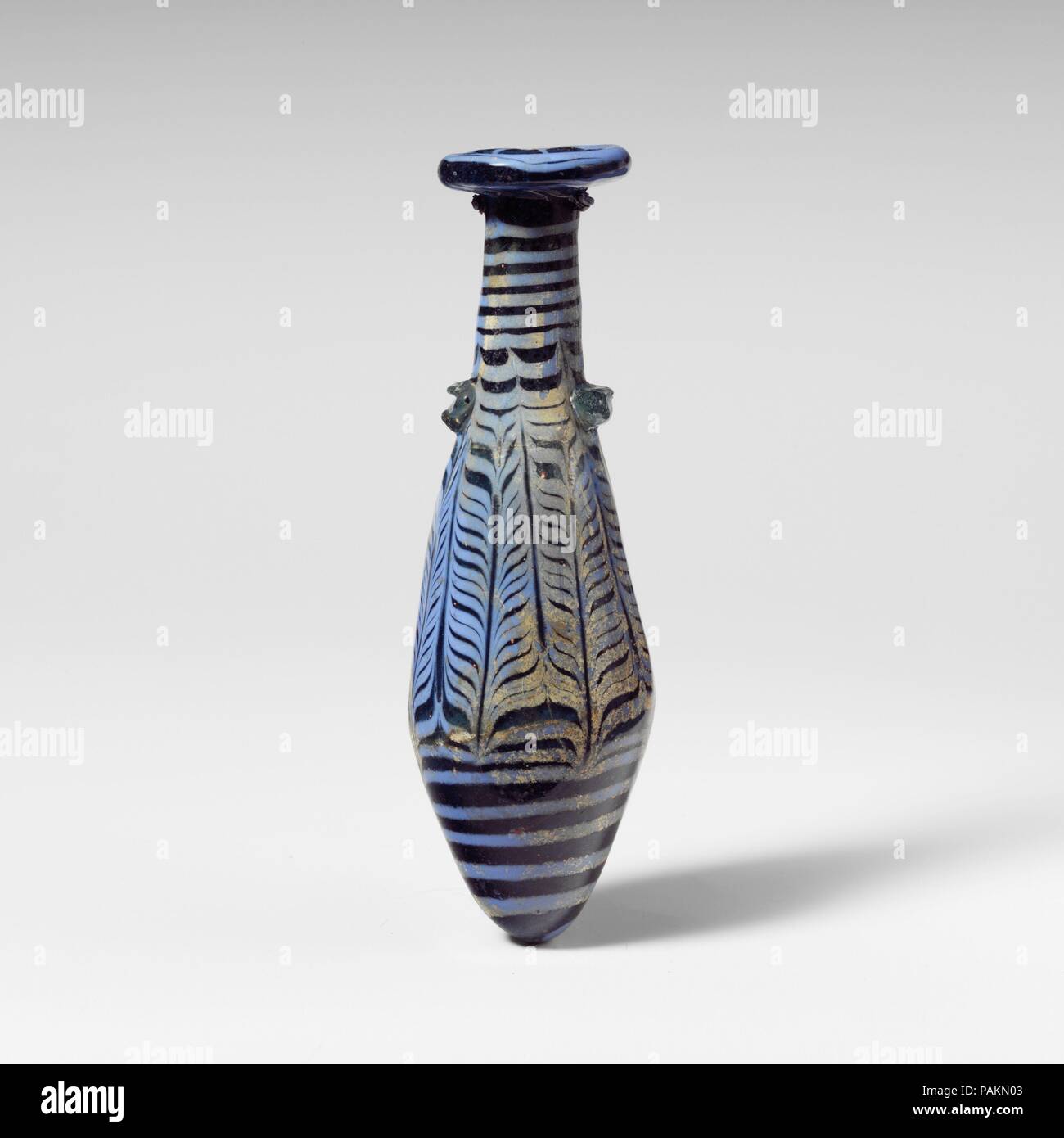 Glass alabastron (perfume bottle). Culture: Greek, Eastern Mediterranean. Dimensions: H.: 5 in. (12.7 cm). Date: 2nd-1st century B.C..  Translucent blue, with handles in light blue green; trail in opaque greyish light blue.  Broad slightly inward-sloping rim-disk with thick rounded edge and radiating tooling marks on lower surface; tall cylindrical neck, expanding downward; straight-sided fusiform body expanding downward, then tapering in to pointed bottom; two horizontal lug handles applied over trail at top of body, one with a deep horizontal indent in surface; one small marvered blob of tra Stock Photo