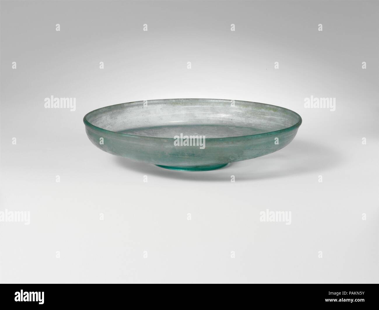 Glass dish. Culture: Roman. Dimensions: H. 1 1/4 in. (3.2 cm)  diameter  5 5/8 in. (14.3 cm). Date: 4th century A.D..  Translucent blue green.  Vertical, slightly thickened, rounded rim; short, convex side to body,  turned in horizontally, and then sloping inwards to thick, flat bottom with upward kick inside and central pontil scar, surrounded by tubular base ring.   Intact; many pinprick bubbles and blowing striations; dulling and faint iridescence. Museum: Metropolitan Museum of Art, New York, USA. Stock Photo