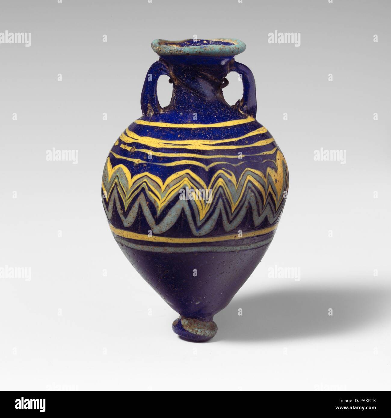 Glass amphoriskos (perfume bottle). Culture: Greek, Eastern Mediterranean. Dimensions: H.: 3 1/2 in. (8.9 cm). Date: late 6th-5th century B.C..  Translucent cobalt blue, with handles in same color; trails in opaque yellow and opaque turquoise blue.  Uneven inward-sloping rim-disk; cylindrical neck with spiral tooling marks, tapering downwards; broad sloping shoulder; top-shaped body; circular base-knob with indent on uneven bottom; two strap handles applied to top of shoulder, drawn up, and pressed onto neck.  Turquoise blue trail attached at edge of rim-disk; a yellow trail applied in an  irr Stock Photo