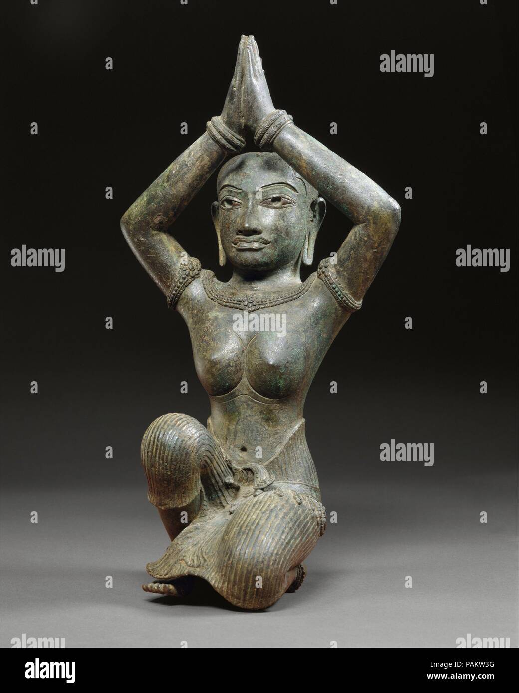 Kneeling Female. Culture: Cambodia. Dimensions: H. 17 (43.2 cm); W. (at knees) 7 3/4 in. (19.7 cm). Date: second half of the 11th century.  This figure, perhaps a Khmer queen, kneels in a posture of adoration with arms raised above her head and palms pressed together. This pose suggests the presence originally of another statue--a god or god-king whom she was worshiping. There are indications that the figure was originally completely gilded. Her eyes are inlaid with silver, and her pupils and brows are hollowed out to receive an inlay, perhaps of black glass. Museum: Metropolitan Museum of Art Stock Photo