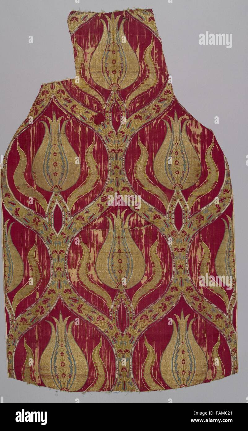 Fragment. Dimensions: Textile: L. 44 in. (111.8 cm)  W. 26 3/4 in. (67.9 cm). Date: 16th century.  Ottoman lampas and velvet textiles often feature large-scale designs displayed within the ogival framework. This example incorporates an elongated tulip flanked by two wavy lines and divided by saz style serrated leaves centering tiny blossoms. Delicate carnations and tulips wind their way along vines inside the ogival lattice. Though slightly worn, the gold surface features metal-wrapped thread emphasized by the bright red background, a color combination popular at the Ottoman court. Based on th Stock Photo