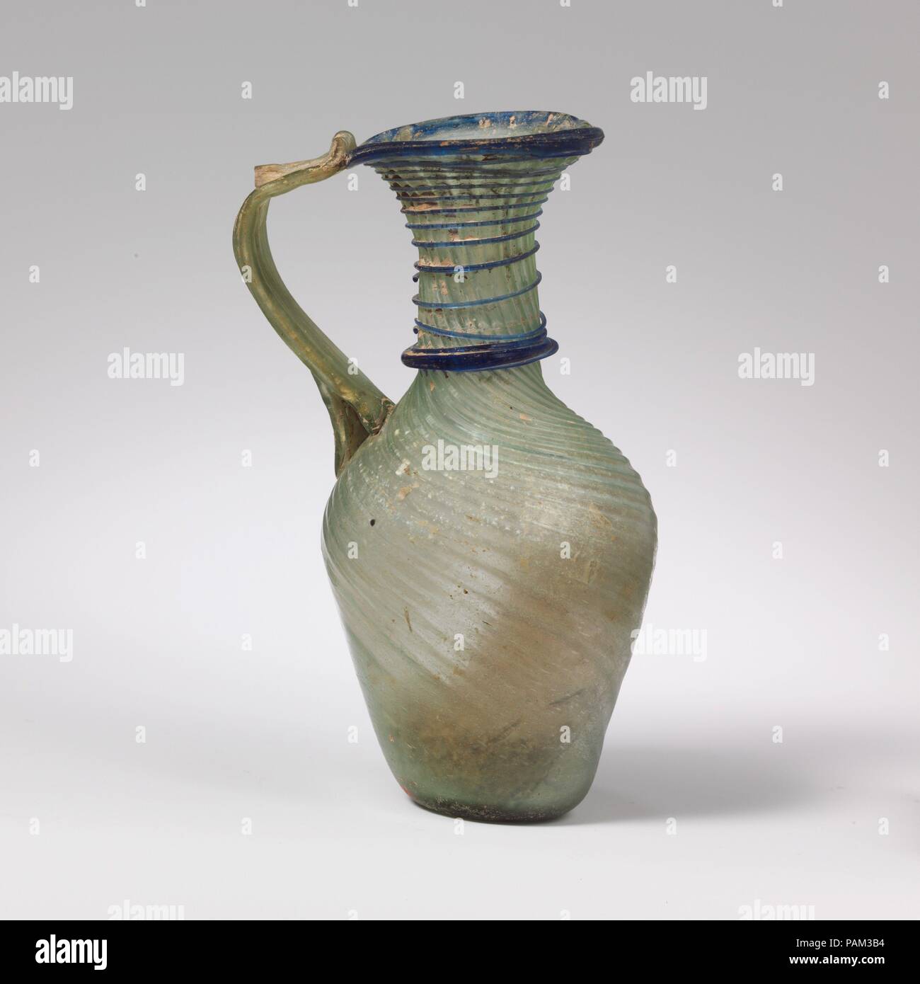 Glass jug. Culture: Roman, Syrian. Dimensions: H.: 4 11/16 in. (11.9 cm). Date: 4th-6th century A.D..  Translucent blue green; handle in same color; trails in translucent cobalt blue.  Plain rim, with downward flange on one side; flaring mouth; cylindrical neck, expanding slightly downwards; slanting, rounded shoulder; funnel-shaped body;  thick bottom with kick and small, central pontil scar; reeded strap handle attached unevenly to shoulder, drawn up and outwards in a curve, then attached to edge of rim and trailed back on itself.  Narrow ribs extend in a tight spiral down from left to right Stock Photo