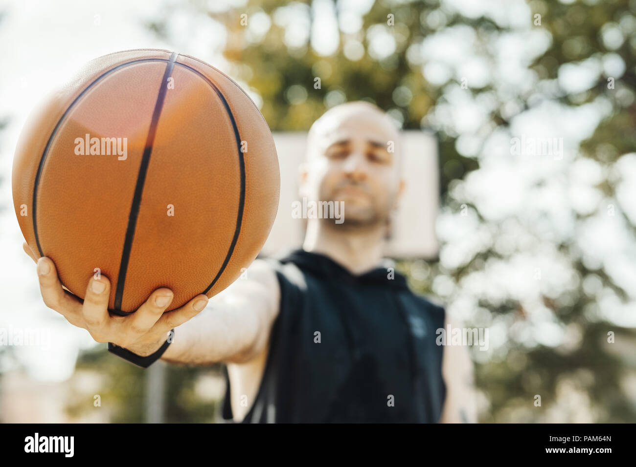 Close up of bald attractive man holding basket ball. Ball is on focus and foreground. Man, basketball hoop and board are on background and blurred. Stock Photo