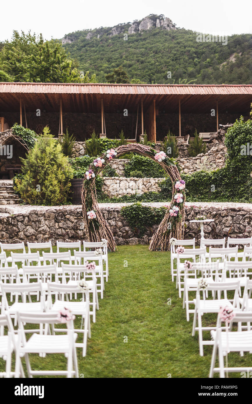 Cozy rustic wedding ceremony with flower arch and wooden white chairs on villa with mountain view Stock Photo