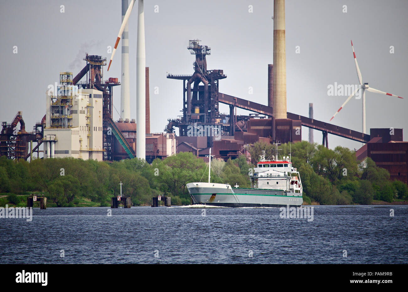 Seagoing cargo ship on a river with green foliage, huge steel mill and several wind power stations in the background Stock Photo