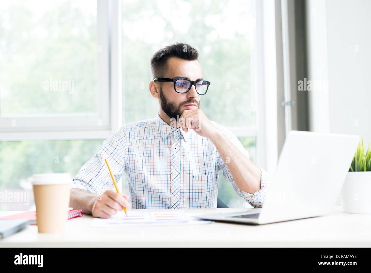Portrait of handsome bearded businessman working at desk in office thoughtfully looking at laptop screen, copy space Stock Photo