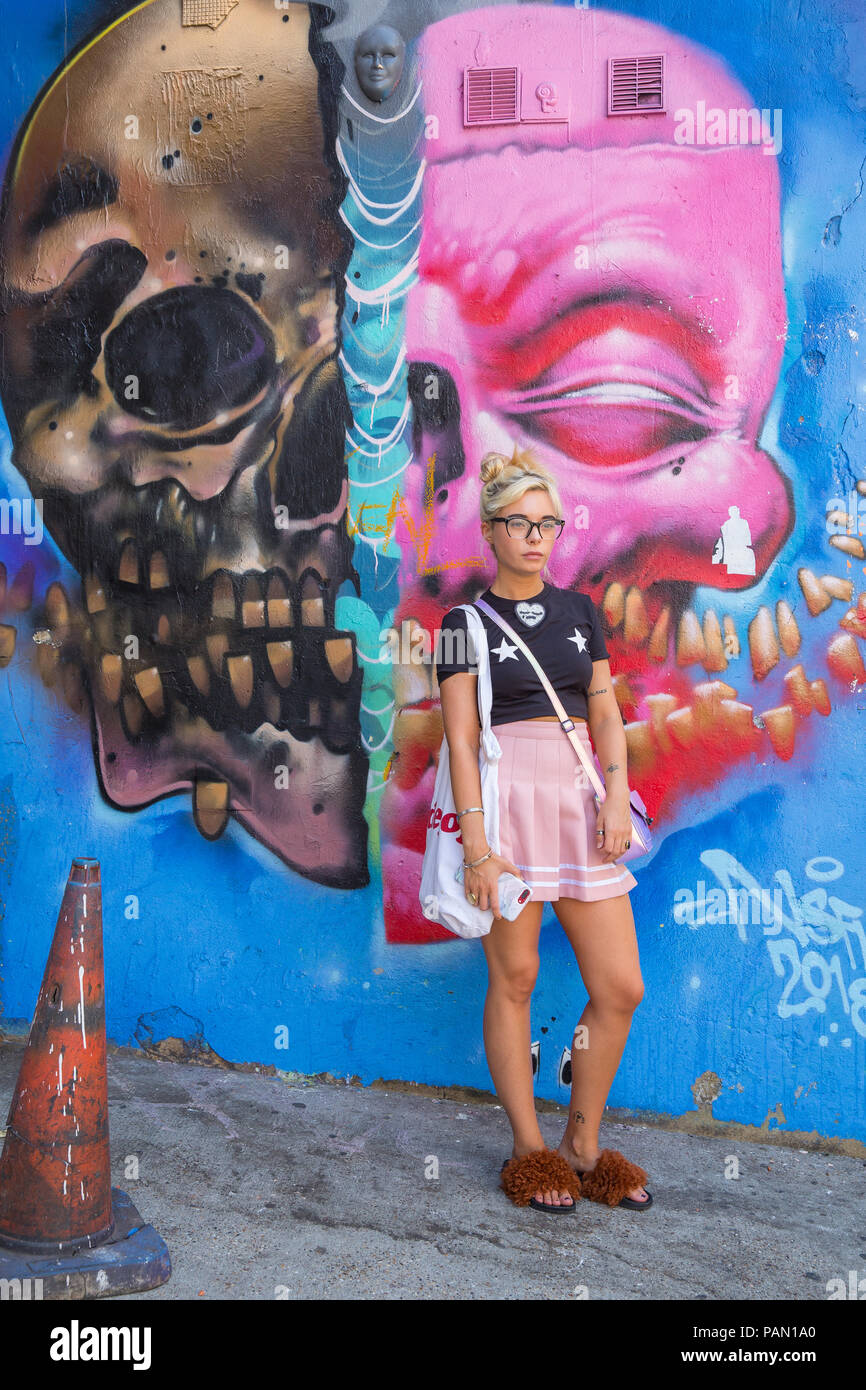 Cool Blond Hip, girl wearing a pink skirt and black short sleeved t-shirt in front of a large graffiti wall on Brick lane in Shoreditch, London, UK. Stock Photo
