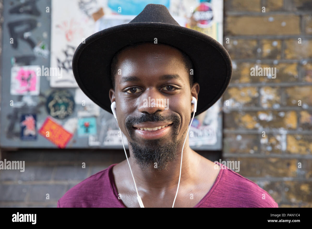 Cool, Hip, hipster, black man wearing a wide brimmed hat and white ear phones in front of a brick wall on Brick lane in Shoreditch, London, UK. Stock Photo