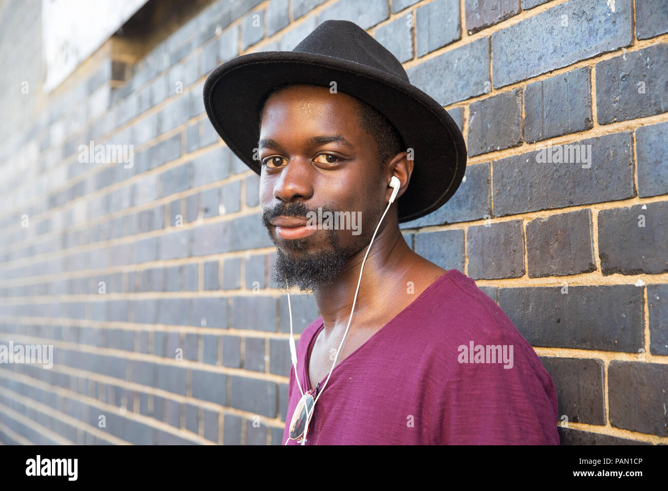 A cool Hipster, black man wearing a wide brimmed hat, purple t-shirt and ear phones, leaning against a wall in Brick Lane, Shoreditch, London. Stock Photo
