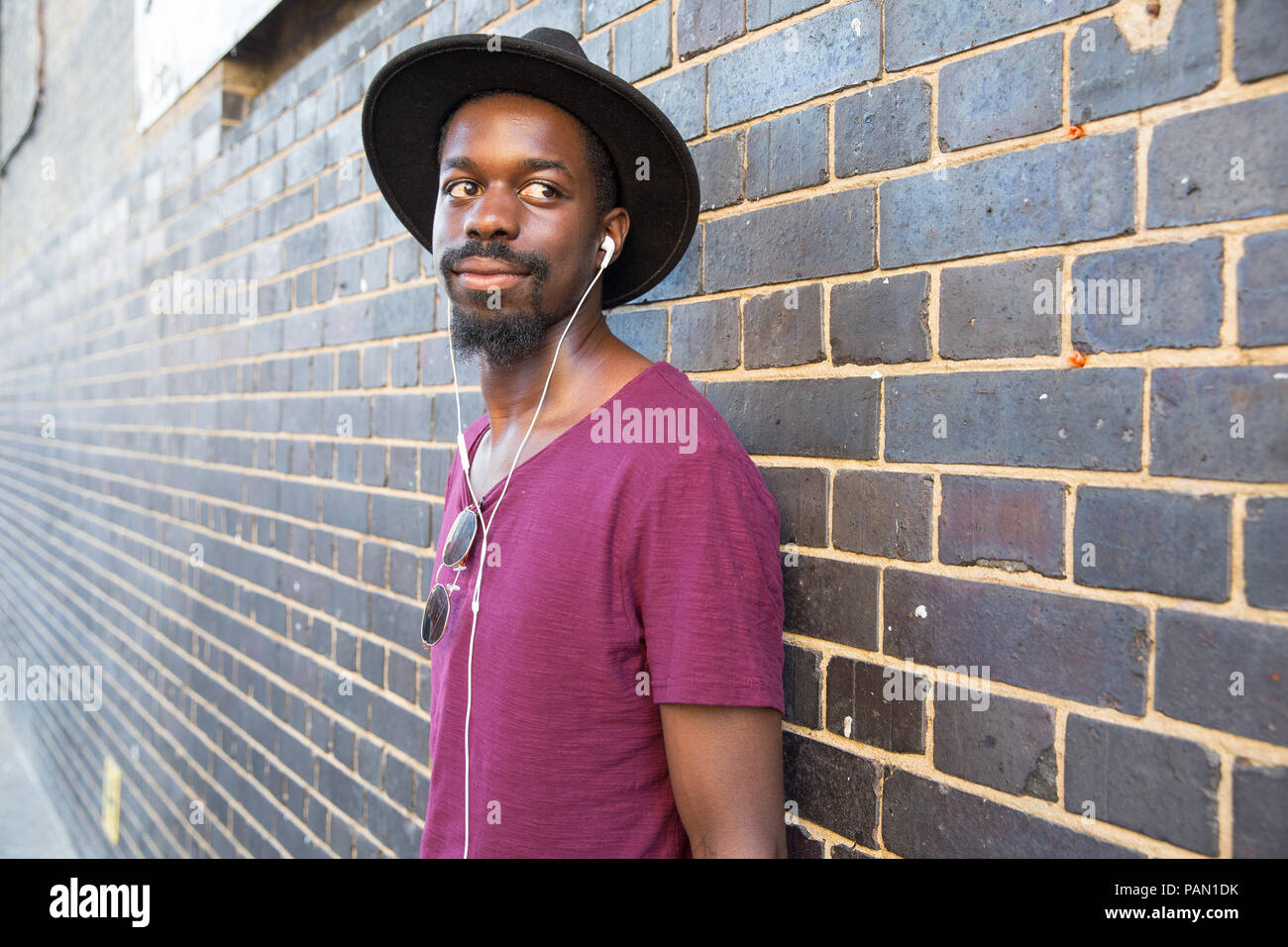 A cool Hipster, black man wearing a wide brimmed hat, purple t-shirt and  ear phones, leaning against a wall in Brick Lane, Shoreditch, London Stock  Photo - Alamy
