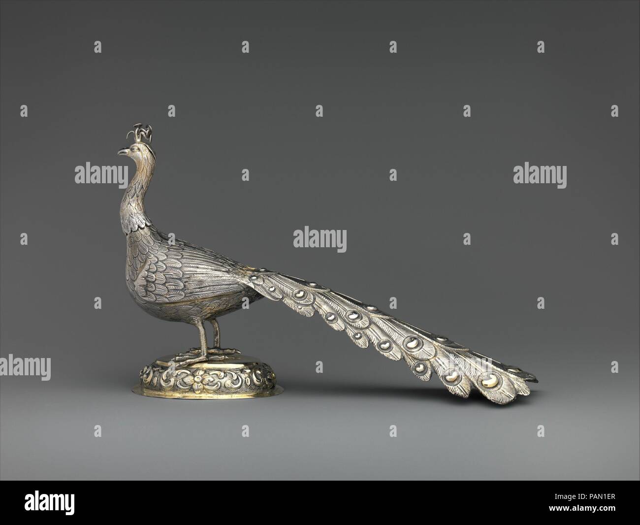 Table decoration in the form of a peacock. Culture: Hungarian, Munkács. Dimensions: Overall: 7 3/16 x 12 in. (18.3 x 30.5 cm). Date: 1787.  The peacock stands with its head slightly tilted, mischievously observing his surroundings. The artist has capture in silver the temperament and characteristic posture of a mature male peacock, an extremely territorial bird. The object's plain oval base, with its convex frame decorated with Baroque flowers and scrolled leaves, is in sharp contrast to the combination of a terrain plinth and stylized Neoclassical garlands on the stands of the pair of peacock Stock Photo
