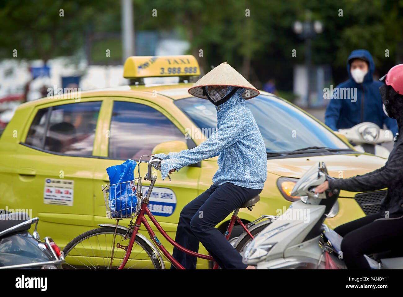 A cyclist in Hanoi, Vietnam, wearing traditional conical straw hat amongst heavy traffic. The iconic hat is worn mostly by women in the Northern part  Stock Photo