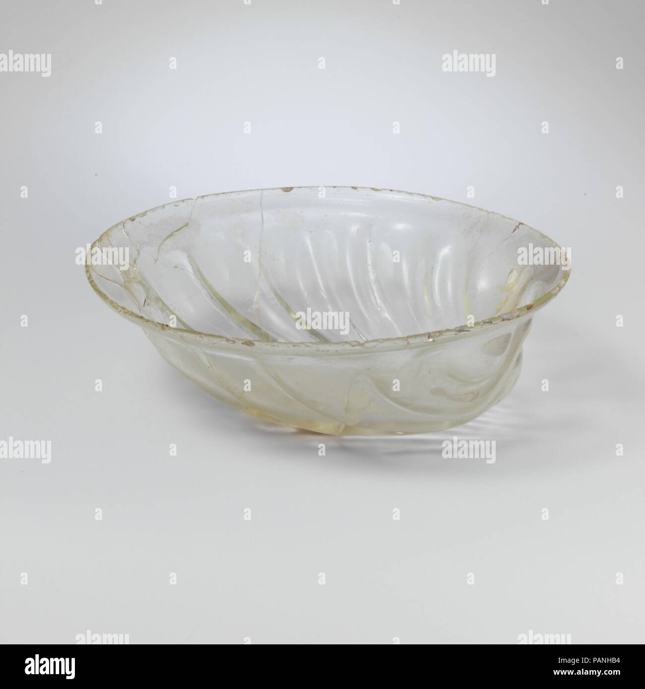 Glass bowl in the form of a shell. Culture: Roman. Dimensions: Diam.: 5 in. (12.7 cm). Date: 1st half of 4th century A.D..  Colorless with pale green tinge.  Flaring, knocked-off rim with shallow S-shaped collar below; body with circular circumference and rounded bottom.  Body formed into the shape of a marine bivalve mollusc (scallop) in relief with eleven ribs radiating from a deep ocellus or umbone in a fan pattern ; ribs become broader and have rounded ends below collar around rim; the ocellus is flanked to either side by a hemispherical  bulge. Below the rim is a faint wheel-abraded line. Stock Photo