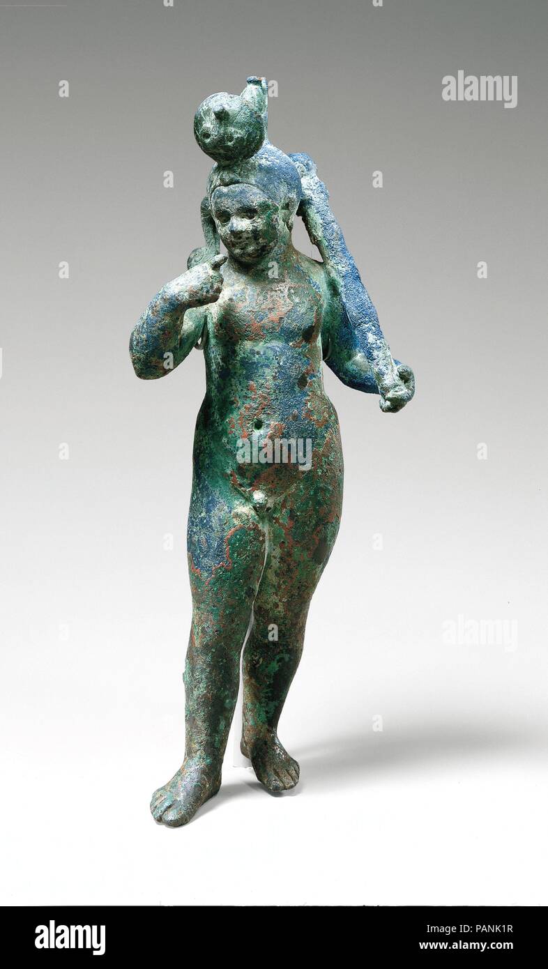 Harpokrates in an Egyptianizing Crown and holding the club of Herakles. Dimensions: H. 18.5 cm. Date: A.D. 1st century.  Harpokrates, or 'Horus the child,' the son of Isis and Osiris/Serapis, is here represented as a chubby toddler with a plump, crooked forefinger reaching toward his small mouth. He holds the knobby club of the Greek god Herakles and on his head wears a vestigial Egyptian double crown detailed with striations and dots. The crown is obscured in the front by a very large sun disk bearing a uraeus. A rarely noted deep blue corrosion gives the piece a rich tone.  Harpokrates, the  Stock Photo