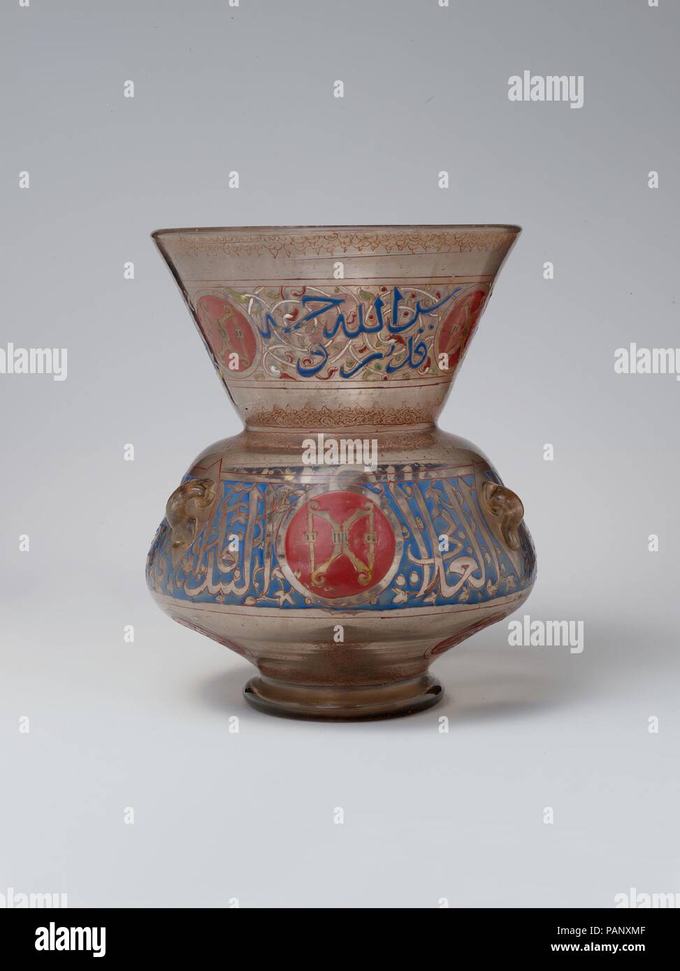 Mosque Lamp for the Mausoleum of Amir Aydakin al-'Ala'i al-Bunduqdar. Dimensions: H. 10 3/8 in. (26.4 cm)  Diam. of rim 8 1/4 in. (21 cm). Date: shortly after 1285.  This lamp's inscriptions reveal that it was ordered for Aidakin's mausoleum (turba), a building still standing in Cairo. Mamluk amirs adopted emblems, often connected with their ceremonial roles at court, which decorated the objects and buildings they commissioned. Here, the motif of two gold crossbows against a red shield illustrates the office of bunduqdar (bow-keeper). Museum: Metropolitan Museum of Art, New York, USA. Stock Photo