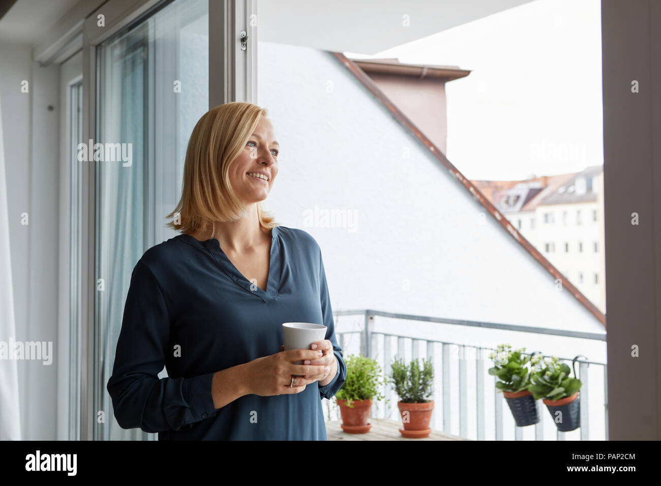 Smiling woman holding cup of coffee looking out of balcony door Stock Photo