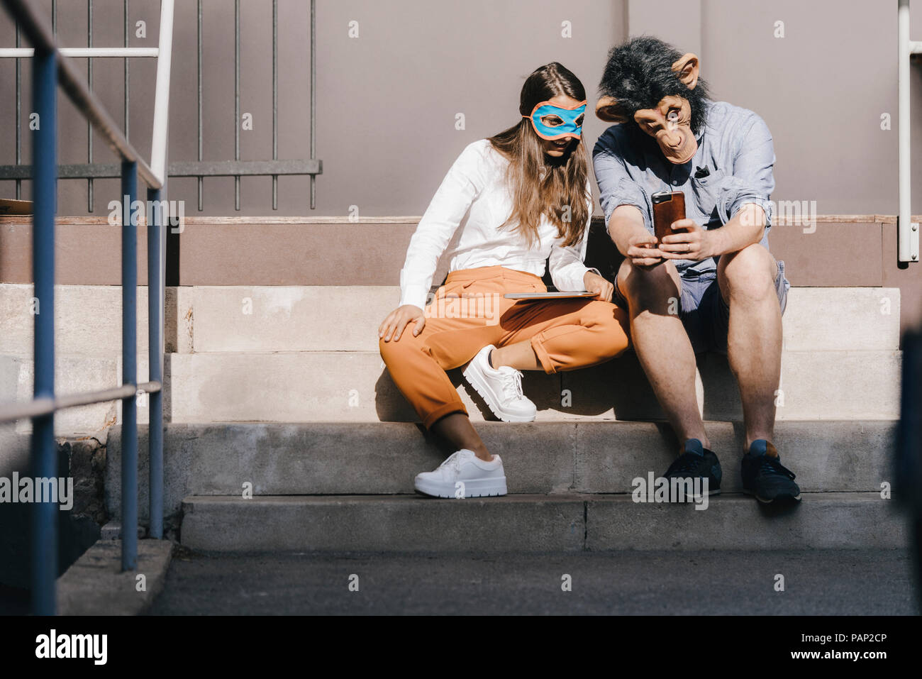 Couple in disguise sitting on stairs, using smartphone Stock Photo