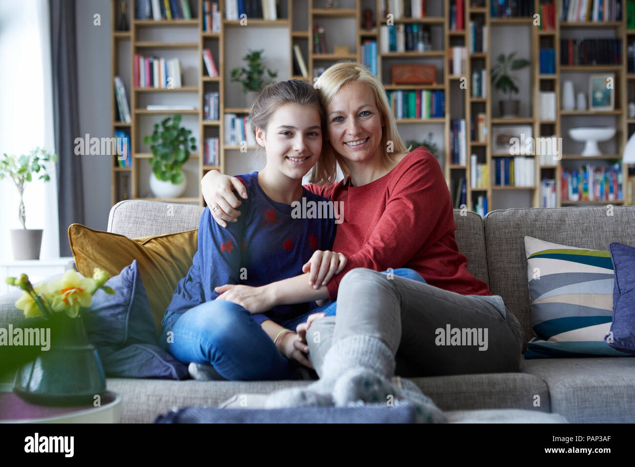 Mother and adolescent daughter sitting on couch with arms around Stock Photo