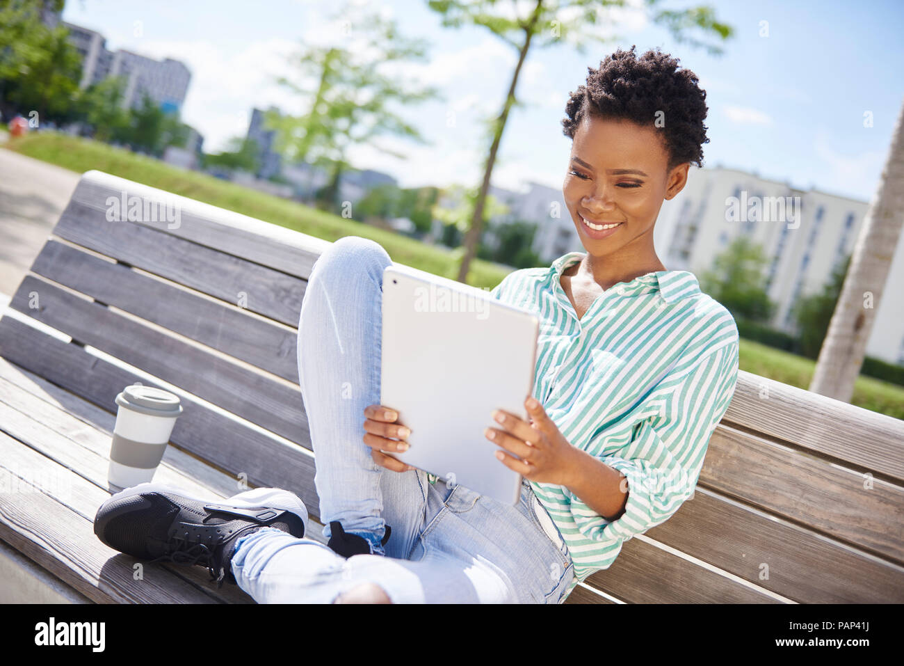 Portrait of smiling young woman with coffee to go sitting on bench looking at tablet Stock Photo
