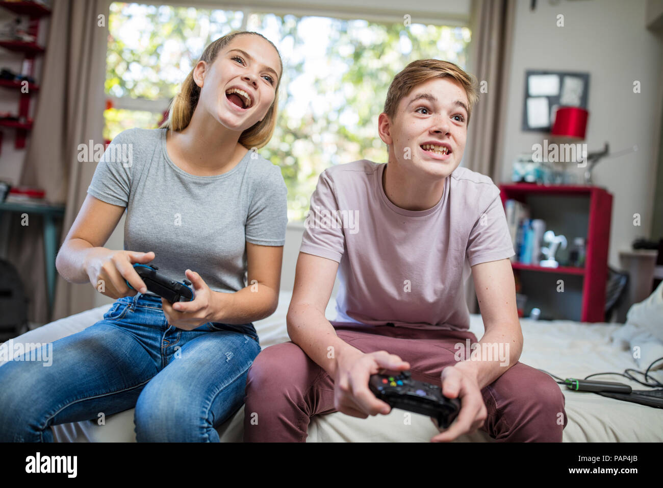 Happy teenage girl and boy sitting on bed playing video game Stock Photo