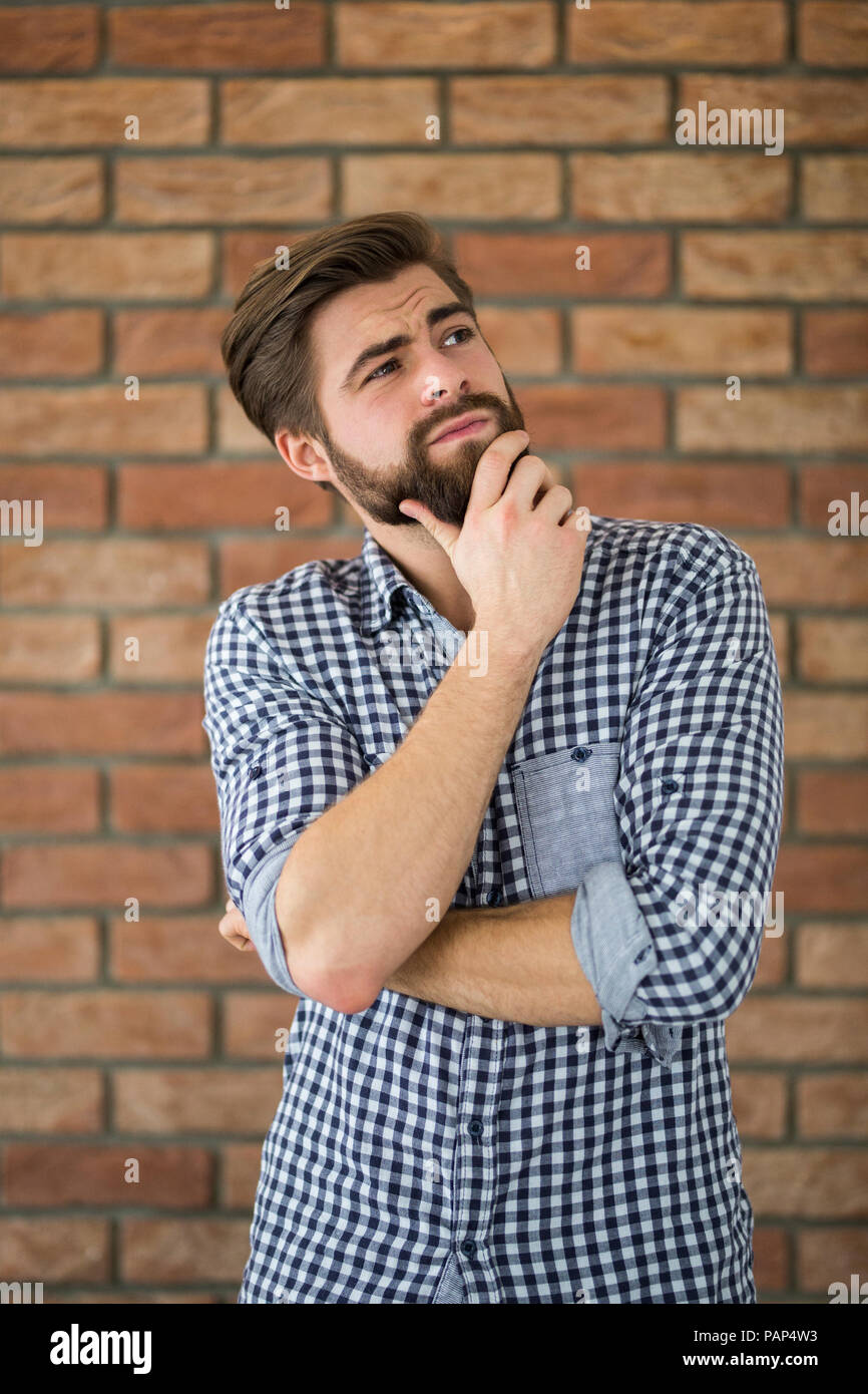 Portrait of bearded young man in front of brick wall thinking Stock Photo