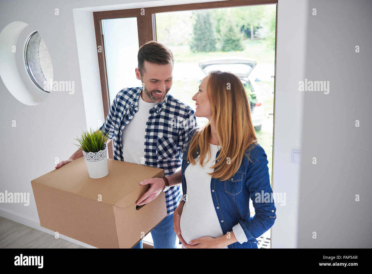 Happy man and pregnant woman moving into new flat carrying cardboard box Stock Photo