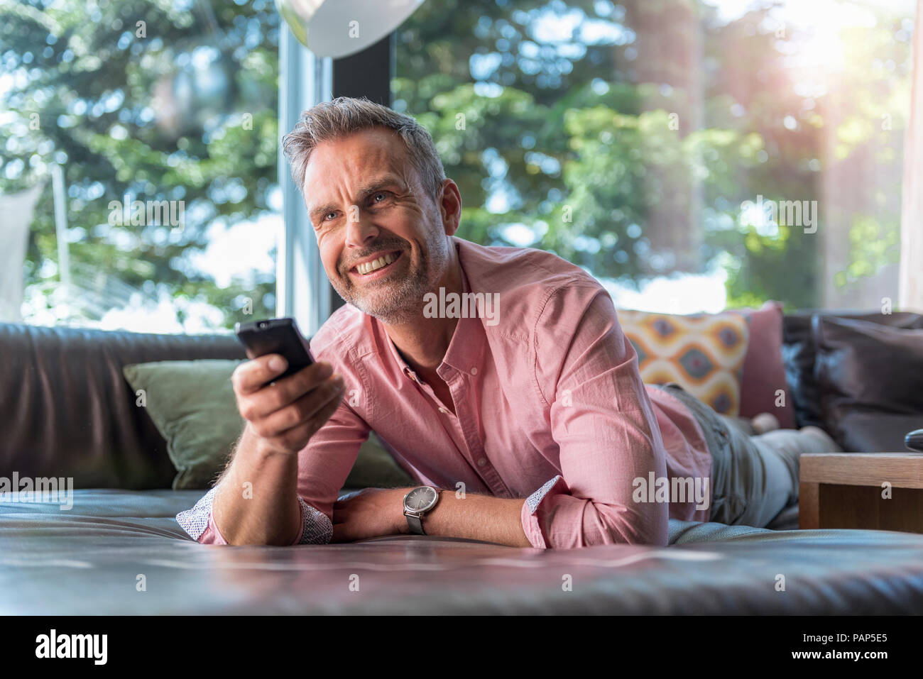Smiling mature man lying on couch at home using remote control Stock Photo