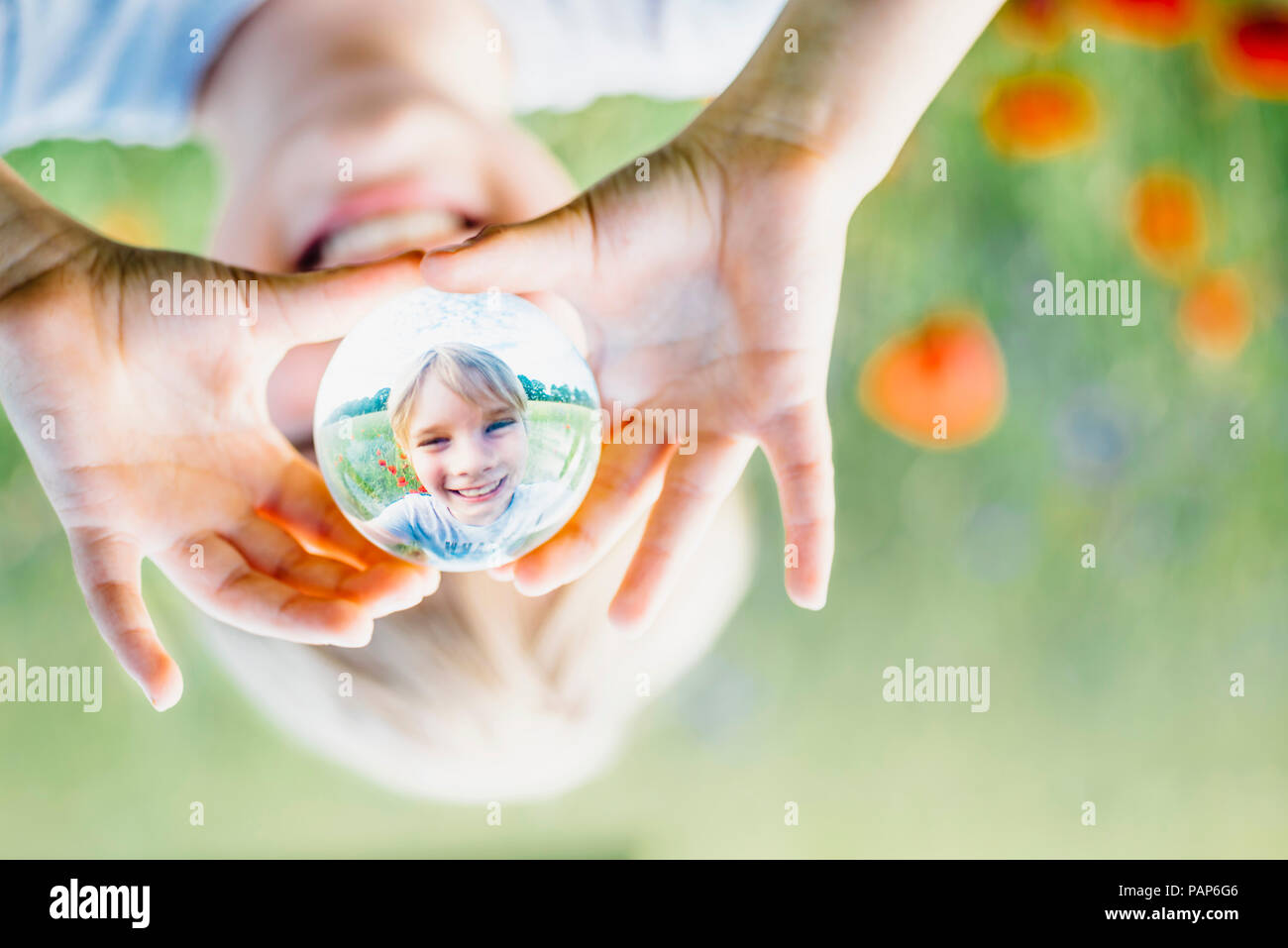 Reflection of smiling boy holding tansparent sphere in poppy field Stock Photo