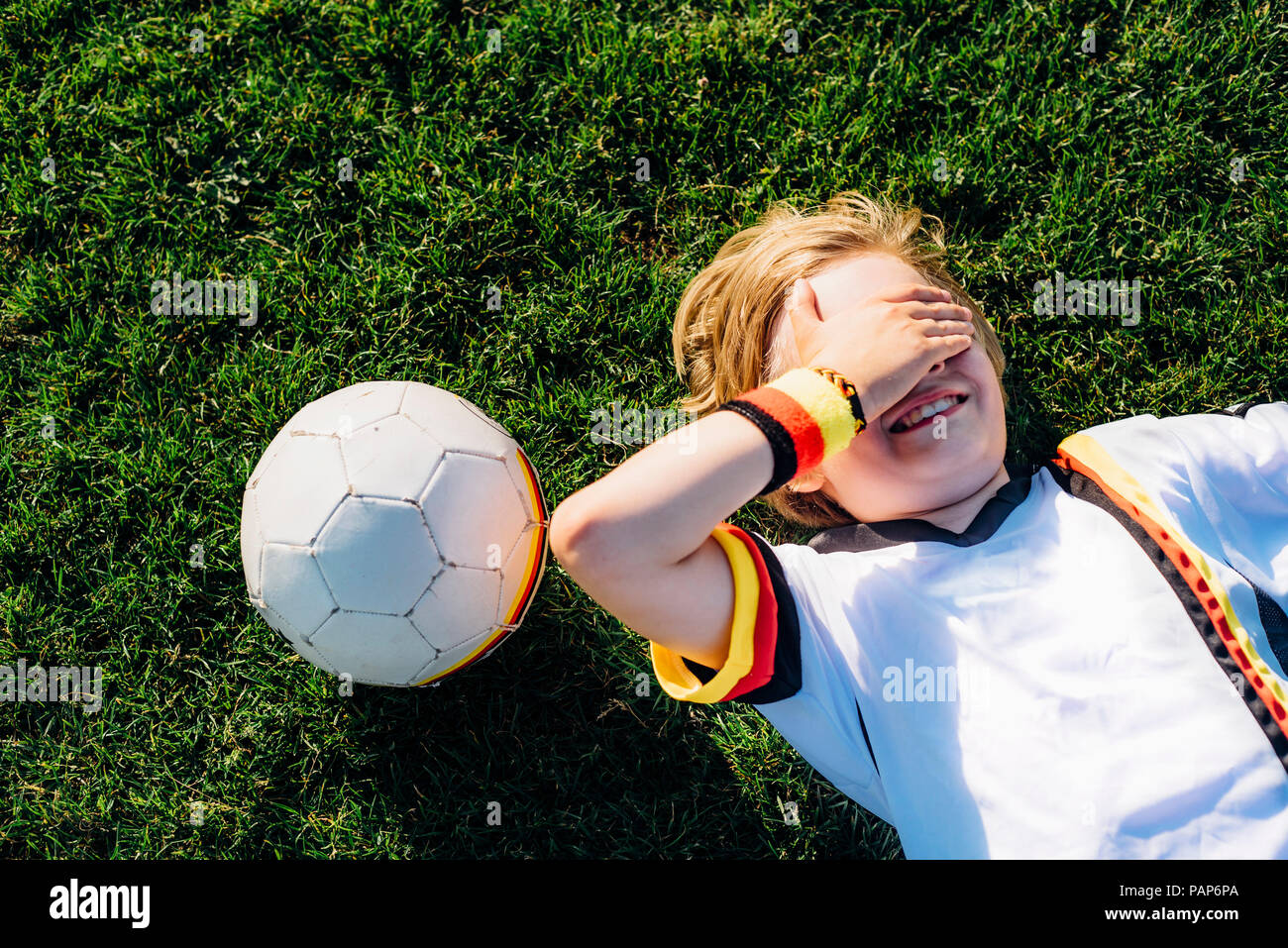 Boy in German soccer shirt lying on grass, covering eyes Stock Photo