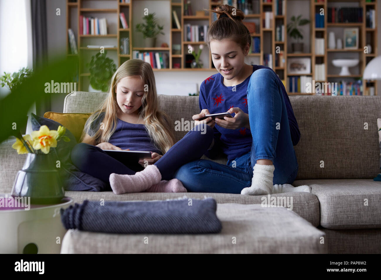 Two sisters sitting on sofa, playing and catting with digital devices Stock Photo