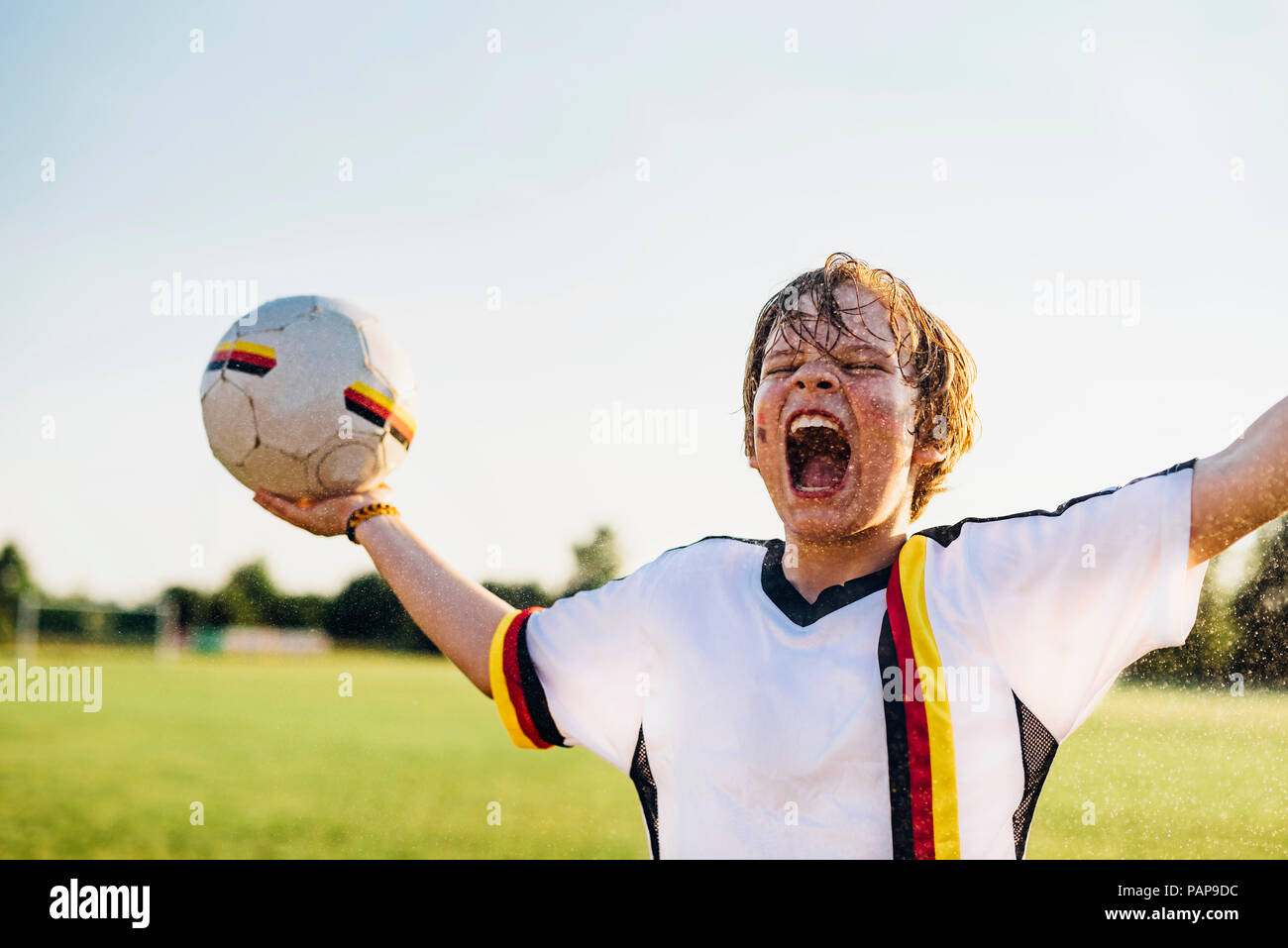 Boy wearing German soccer shirt screaming for joy, standing in water splashes Stock Photo