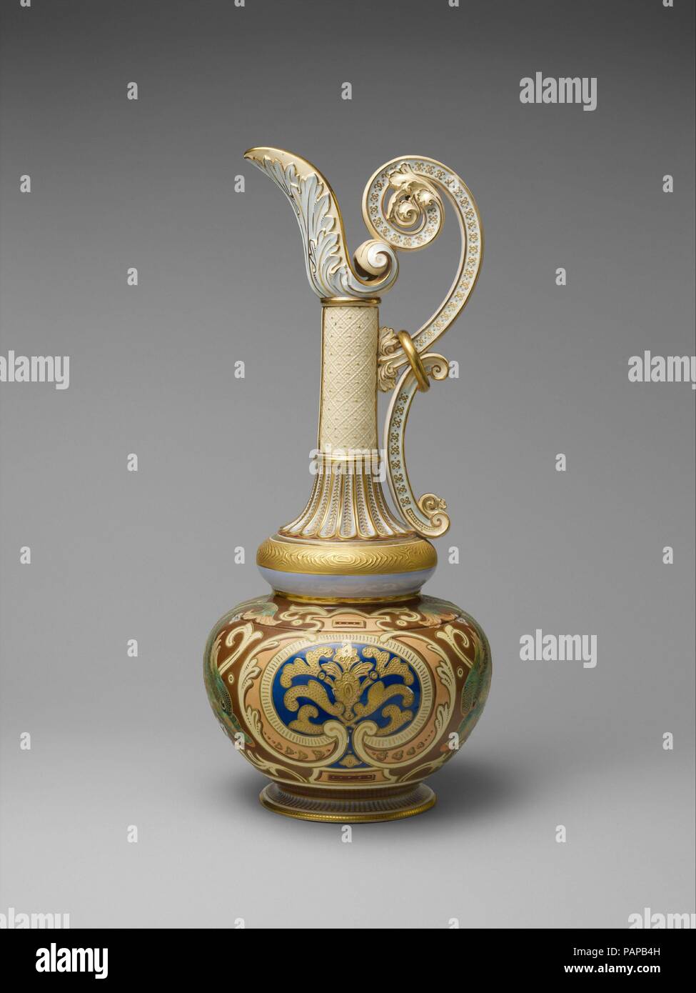 Ewer. Culture: American. Designer: Designed by Edward Lycett (1833-1910). Dimensions: H. 22 in. (55.9 cm). Manufacturer: Faience Manufacturing Company (American, New York, 1881-1892). Date: 1886-90.  At 22 inches high, this ewer is one of the five largest known vessels produced by the Faience Manufacturing Company, and this example displays one of at least four different motifs associated with this form. Rising from a bulbous double-gourd-form body, stacked elements that display classicizing ornament emphasize the ewer's elongated form. Opposing foliate scrolls, joined by an illusionistic gilt Stock Photo
