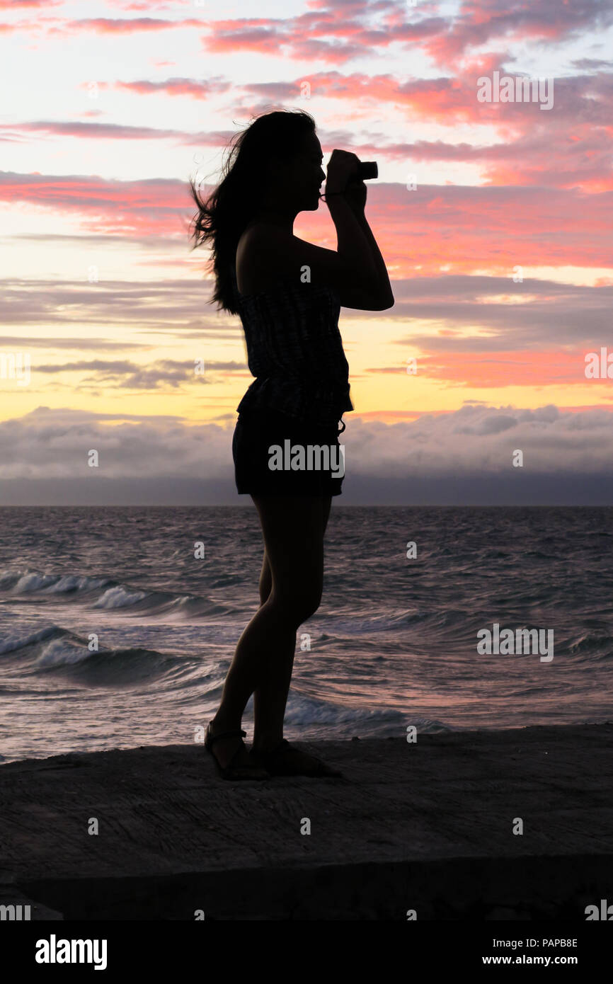 Young Woman Silhouette Profile, Taking Pictures of a stunning pink island sunset - Bohol, Philippines Stock Photo