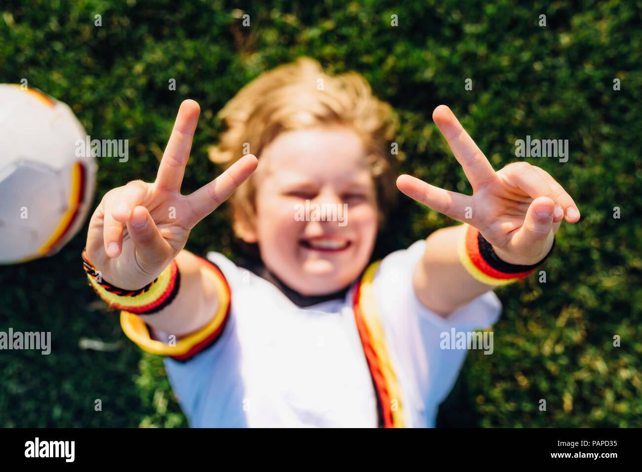 Boy in German soccer shirt lying on grass, making victory sign Stock Photo