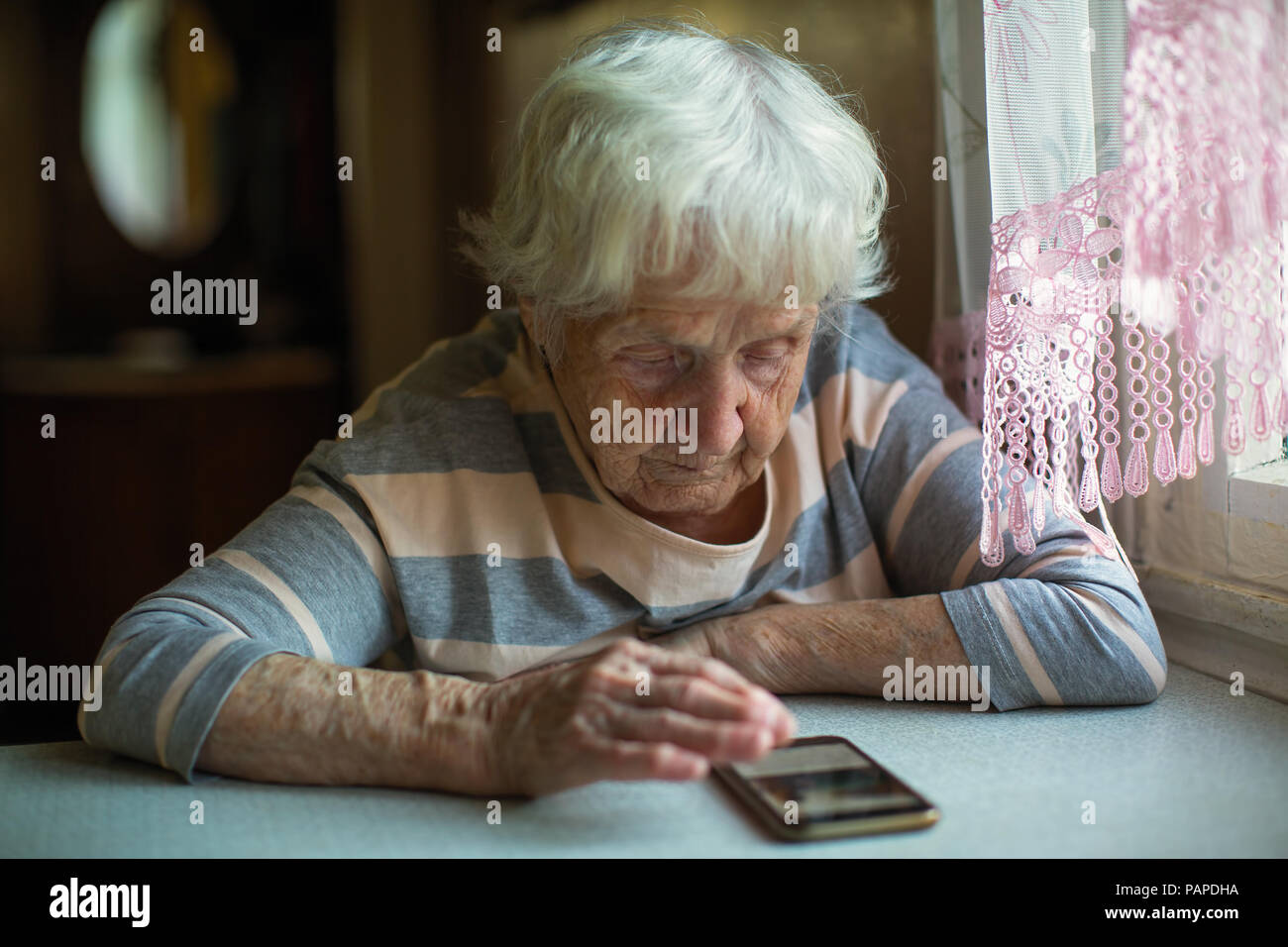 An elderly woman sits with smartphone at the table. Stock Photo