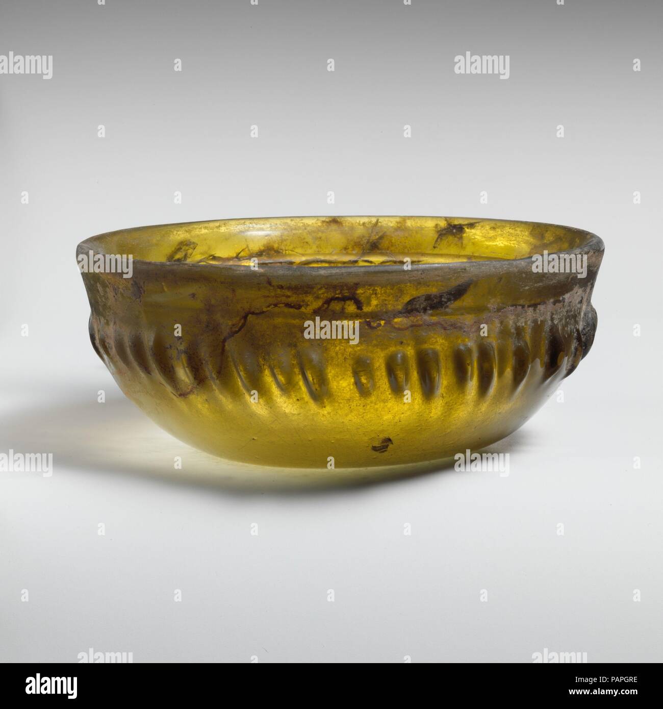 Glass ribbed bowl. Culture: Greek, Eastern Mediterranean. Dimensions: H.: 2 3/16 in. (5.6 cm)  Diam.: 5 3/8 in. (13.7 cm). Date: 1st century B.C..  Translucent yellow green.  Outsplayed rim, with upright rounded edge and plain inward-sloping band below; convex side curving in downwards; concave bottom.  On interior, a single broad horizontal groove at junction of rim and side; on exterior, forty-three short, unevely-spaced ribs of varying length and width, arranged around bulging middle section of body, occasionally flanked by vertical tooling marks.  Intact; some internal strain cracks; some  Stock Photo