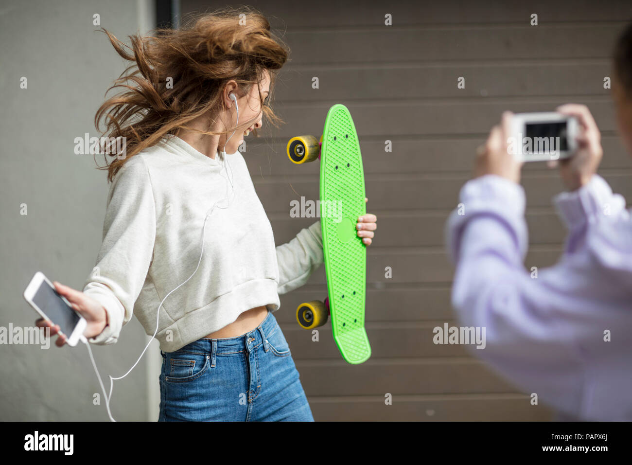 Friend taking picture of carefree teenage girl dancing while holding skateboard and listening to music Stock Photo