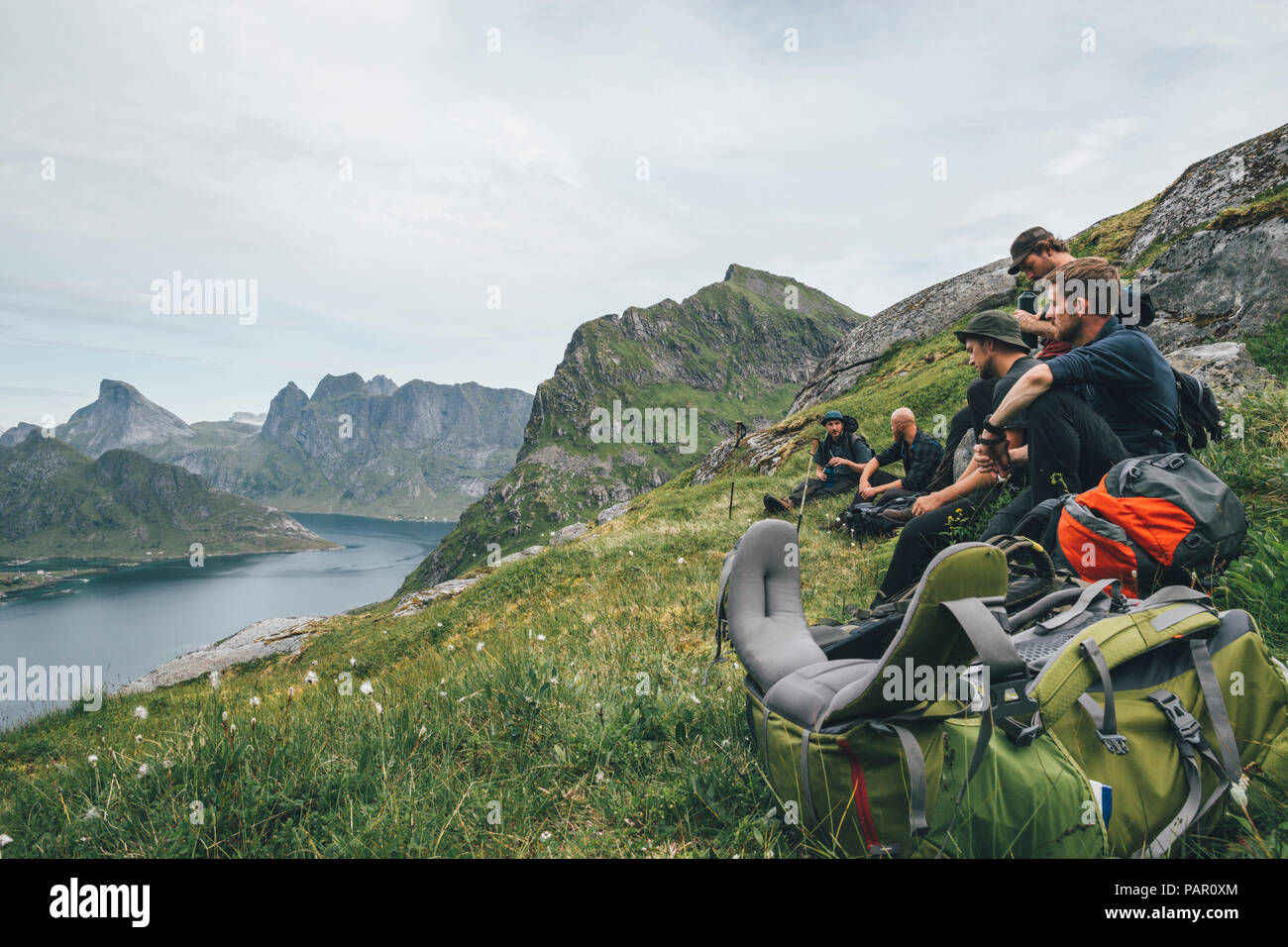 Norway, Lofoten, Moskenesoy, Group of young men sitting on grass, looking over Kjerkefjord Stock Photo
